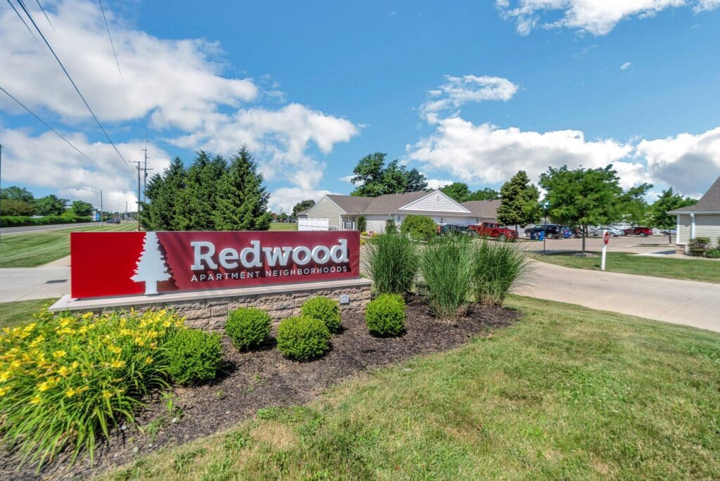 Redwood Apartment Neighborhoods sign surrounded by landscaping, with residential buildings and a driveway in the background under a partly cloudy sky.