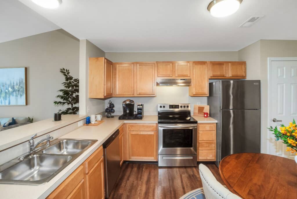 Modern kitchen with light wood cabinets, stainless steel appliances, double sink, and a round dining table with flowers; a living area is partially visible in the background.
