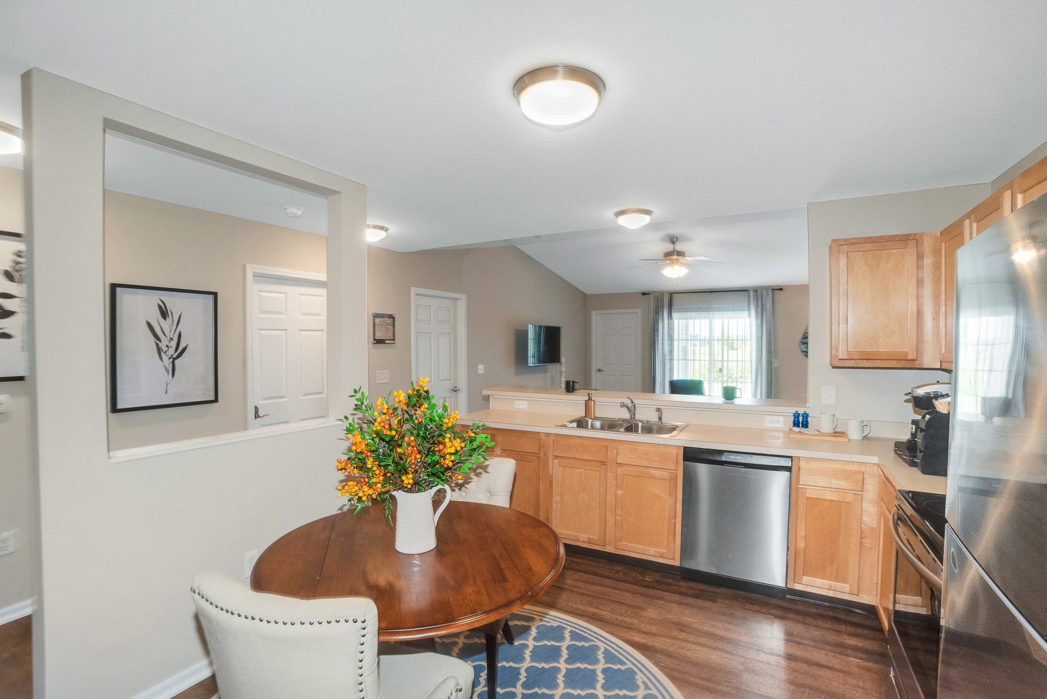 A kitchen and dining area with wood cabinets, stainless steel appliances, a round table with flowers, and a patterned rug on a wood floor.