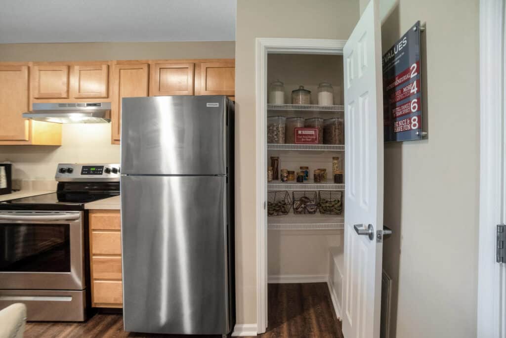 A stainless steel refrigerator and stove are next to an open pantry door with shelves holding jars and containers of food in a kitchen with wooden cabinets.