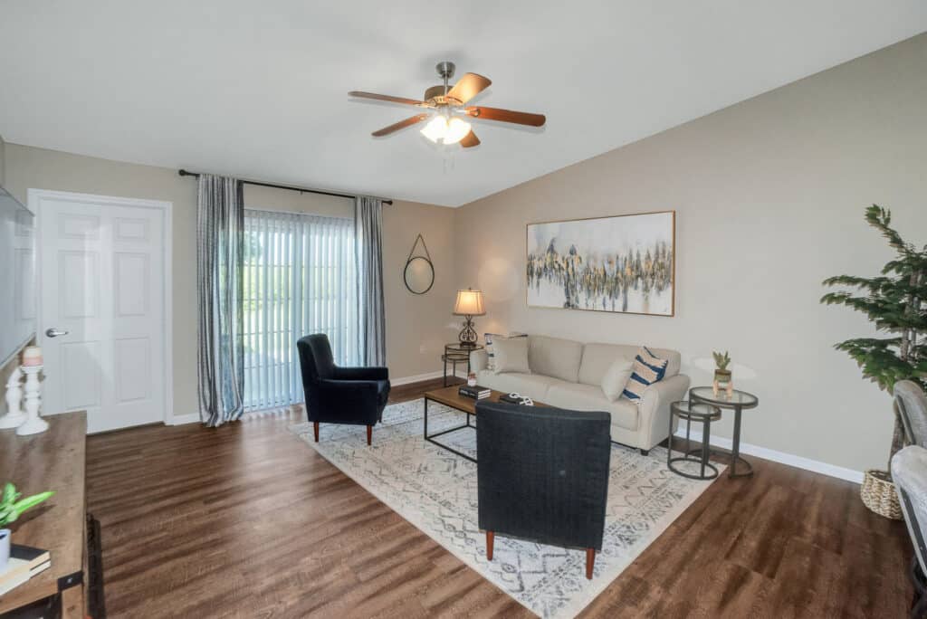 Living room with wood flooring, beige walls, a ceiling fan, a sofa, two black chairs, a rug, a coffee table, a floor lamp, and a large window with sheer curtains.