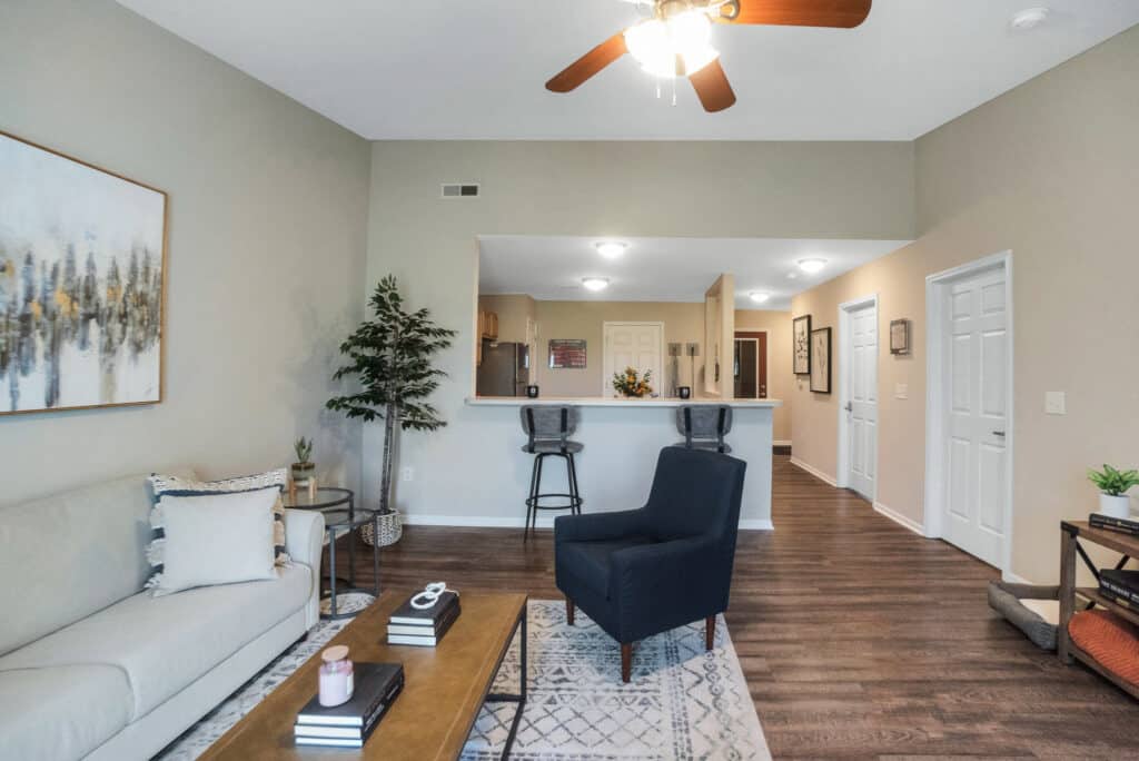 Modern living room with a beige sofa, black armchair, coffee table, barstools at a kitchen counter, artwork on the wall, and a ceiling fan.