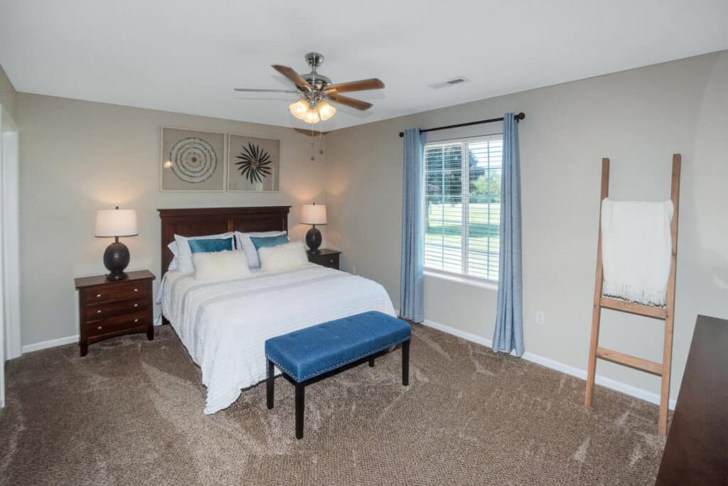 Bedroom with a bed, two nightstands, lamps, a blue bench at the foot of the bed, wall art, a ceiling fan, a window with blinds, and a ladder holding a blanket.