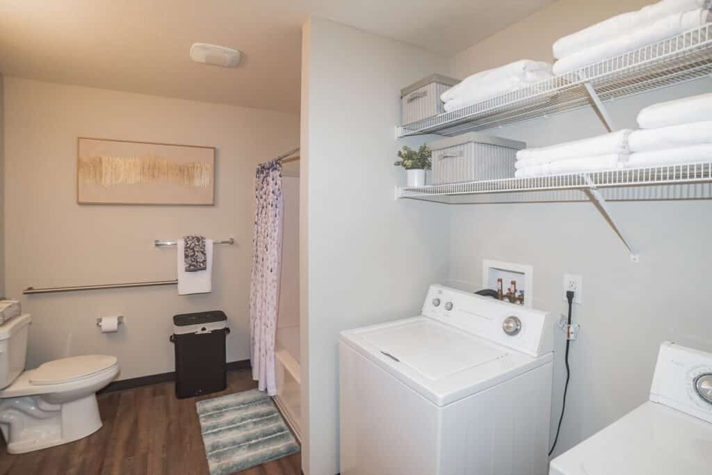 Laundry area with washer and dryer next to open shelves holding folded towels and baskets; bathroom with toilet, shower, and wall art in the background.