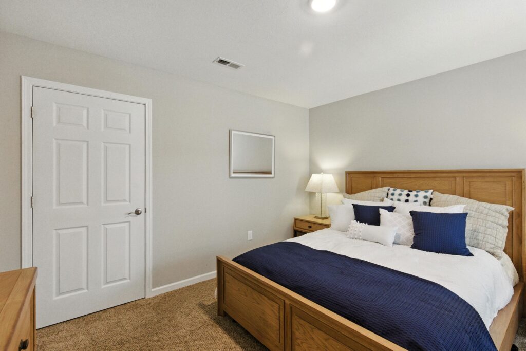 A bedroom with a wooden bed frame, white and navy bedding, a nightstand with a lamp, a closed white door, and a wall-mounted mirror.