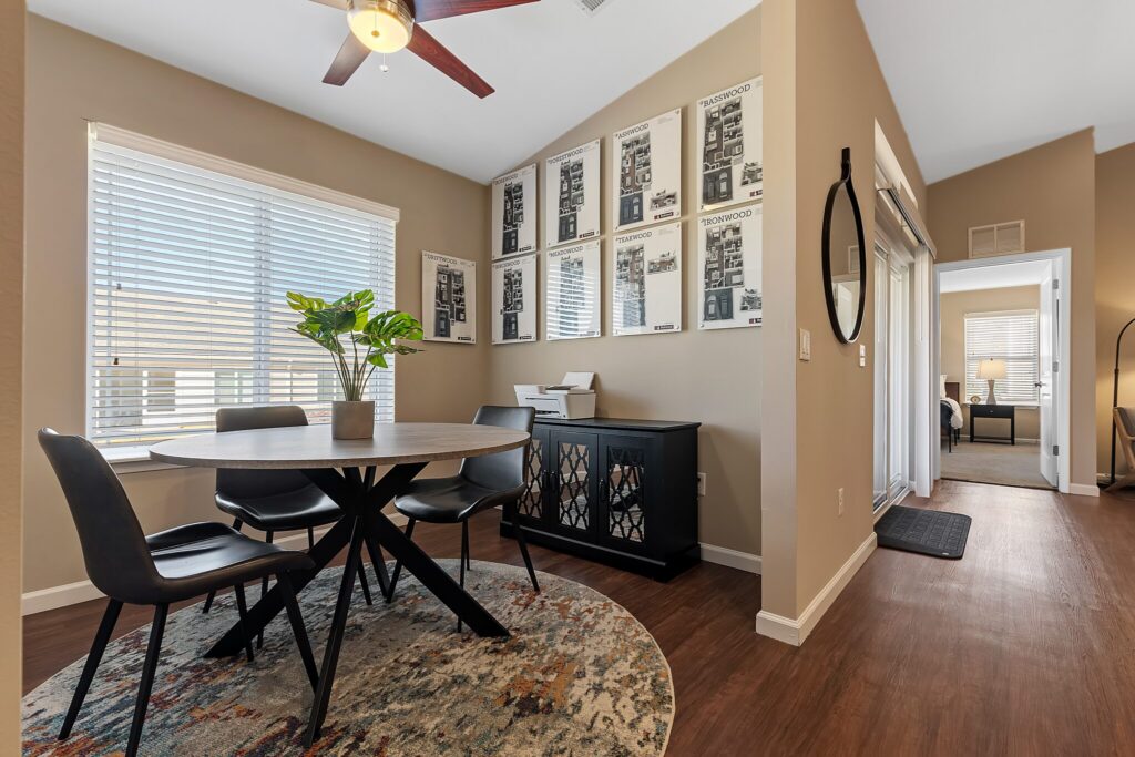 A modern dining area with a round table, four chairs, a plant centerpiece, wall art, wood flooring, and a side cabinet next to a hallway.