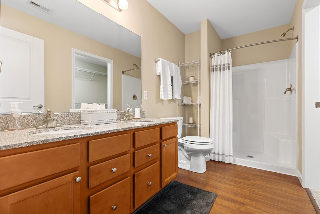 Bathroom with double sink vanity, granite countertop, wood cabinets, white toilet, shower with curtain, towel rack, and wood flooring.