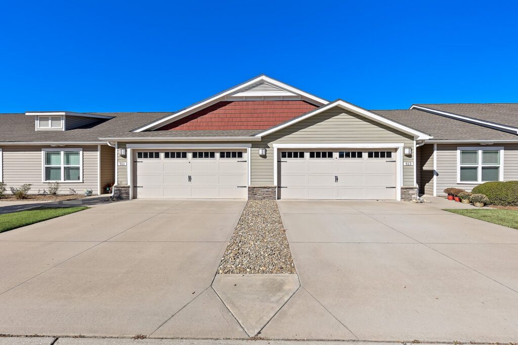 Front view of a modern duplex house with two garage doors, a shared driveway divided by a strip of decorative rocks, and a clear blue sky overhead.