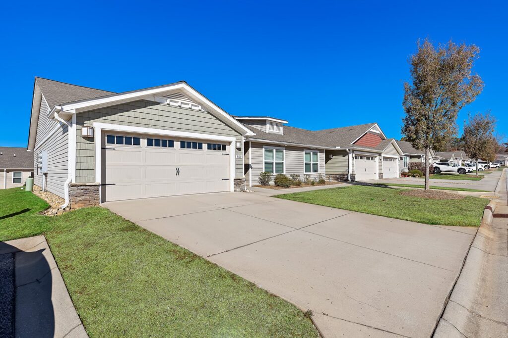 Single-story suburban house with a two-car garage, neatly trimmed lawn, and a clear blue sky overhead on a residential street.