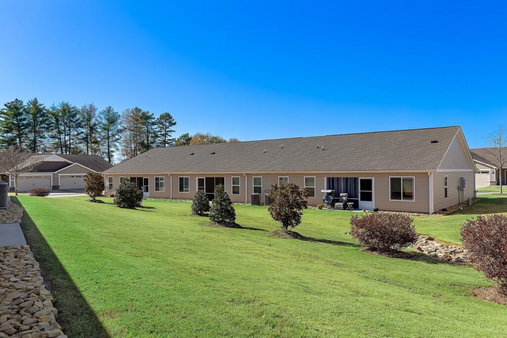 Single-story residential building with multiple units, each with a patio, overlooking a grassy backyard with shrubs and trees under a clear blue sky.