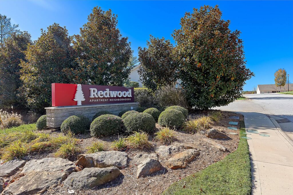 Entrance sign for Redwood Apartment Neighborhoods, surrounded by bushes, rocks, and trees, with a sidewalk and street visible to the right under a clear blue sky.