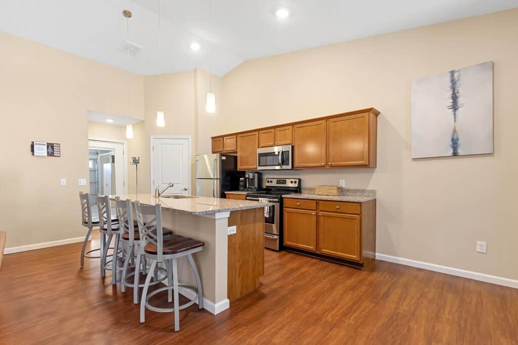 Modern kitchen with stainless steel appliances, wooden cabinets, granite countertops, an island with bar stools, and pendant lighting, set against beige walls and wood flooring.
