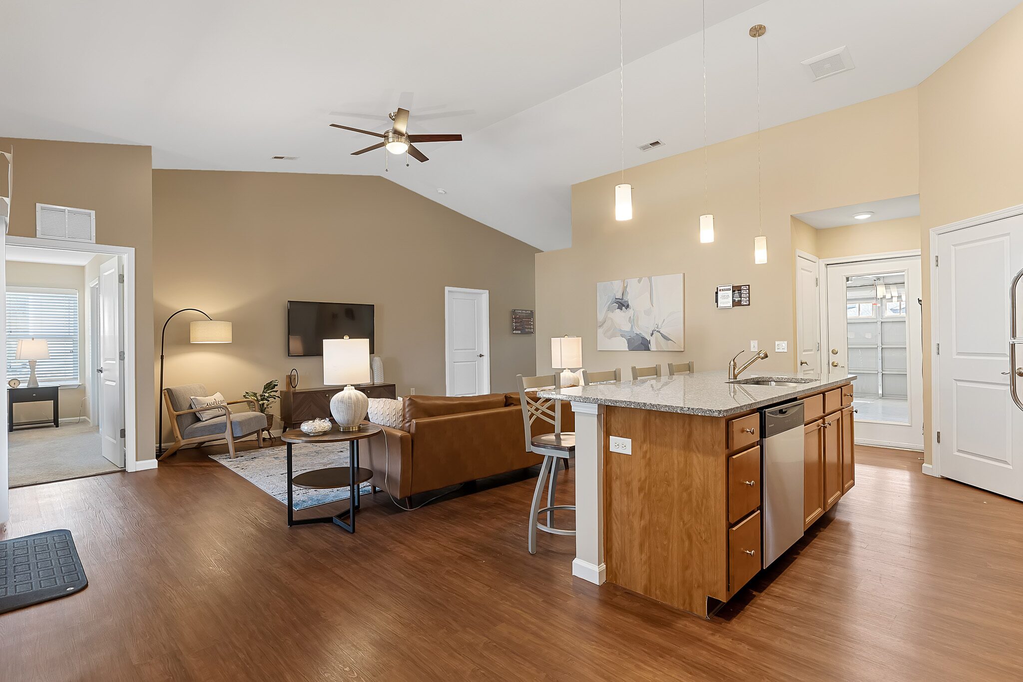 Open-concept living area with a kitchen island, brown cabinets, a leather sofa, TV, and a reading nook; wood flooring and beige walls with modern lighting.