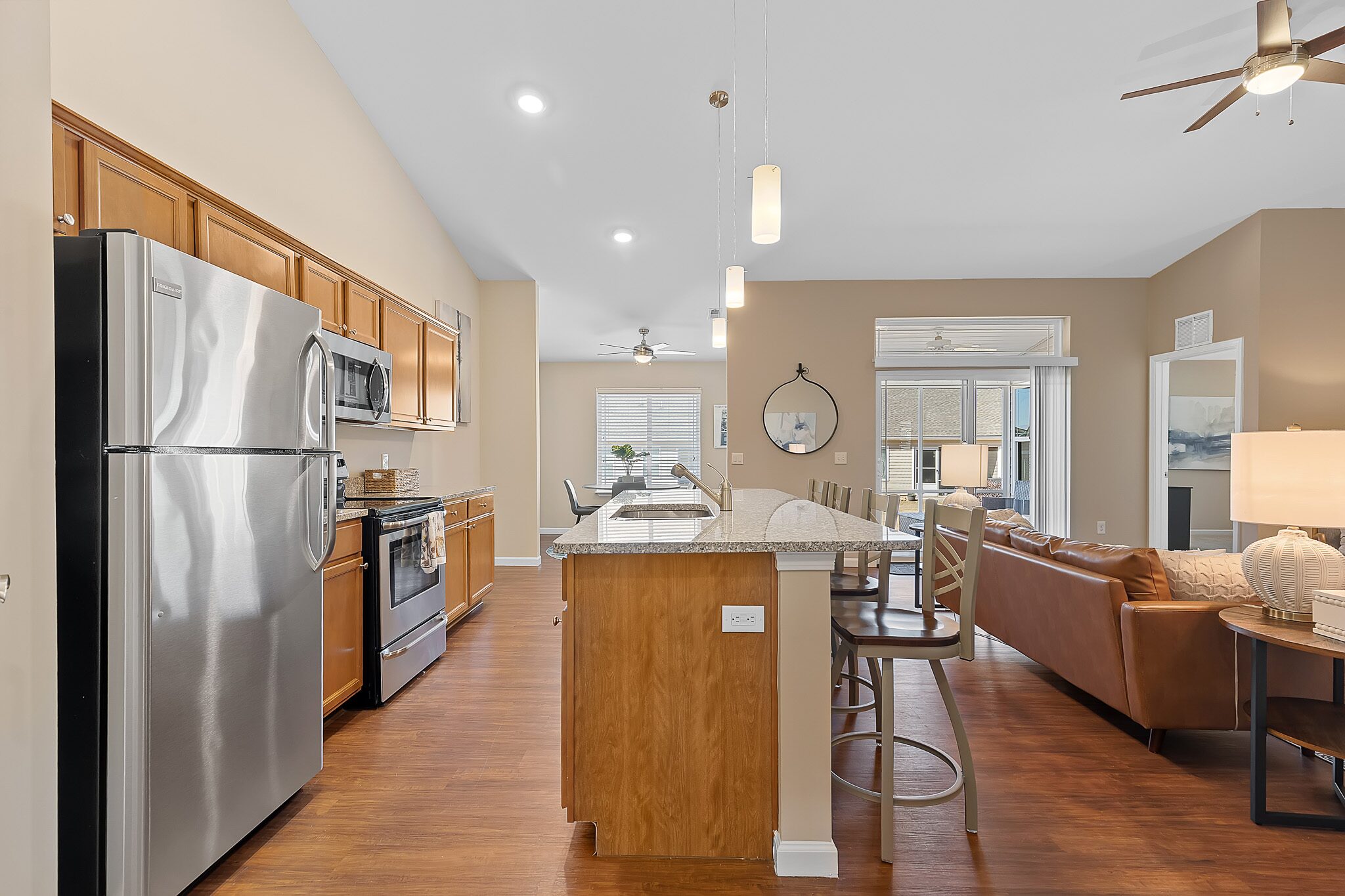Modern open-concept kitchen and living room with stainless steel appliances, wooden cabinets, island with bar stools, and neutral-toned decor.