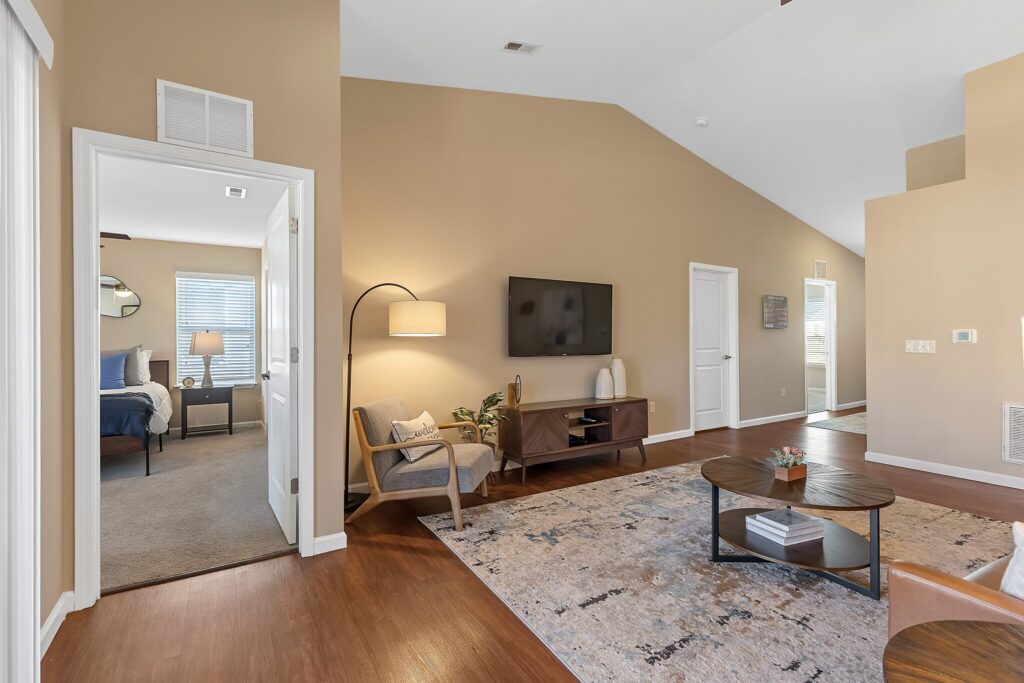 A modern living room with wood flooring, beige walls, a wall-mounted TV, a rug, a coffee table, and doorway leading to a bedroom with a bed and window.
