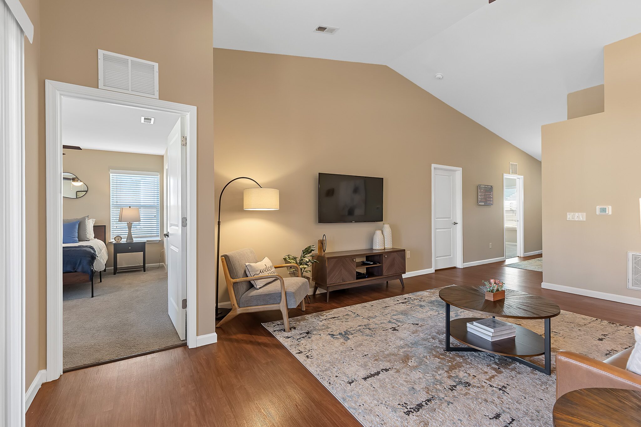 A modern living room with wood flooring, beige walls, a wall-mounted TV, a rug, a coffee table, and doorway leading to a bedroom with a bed and window.