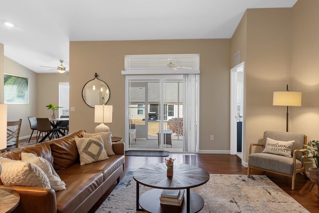 A modern living room with a brown leather sofa, round coffee table, armchair, floor lamp, and glass doors leading to a patio; dining area visible in the background.