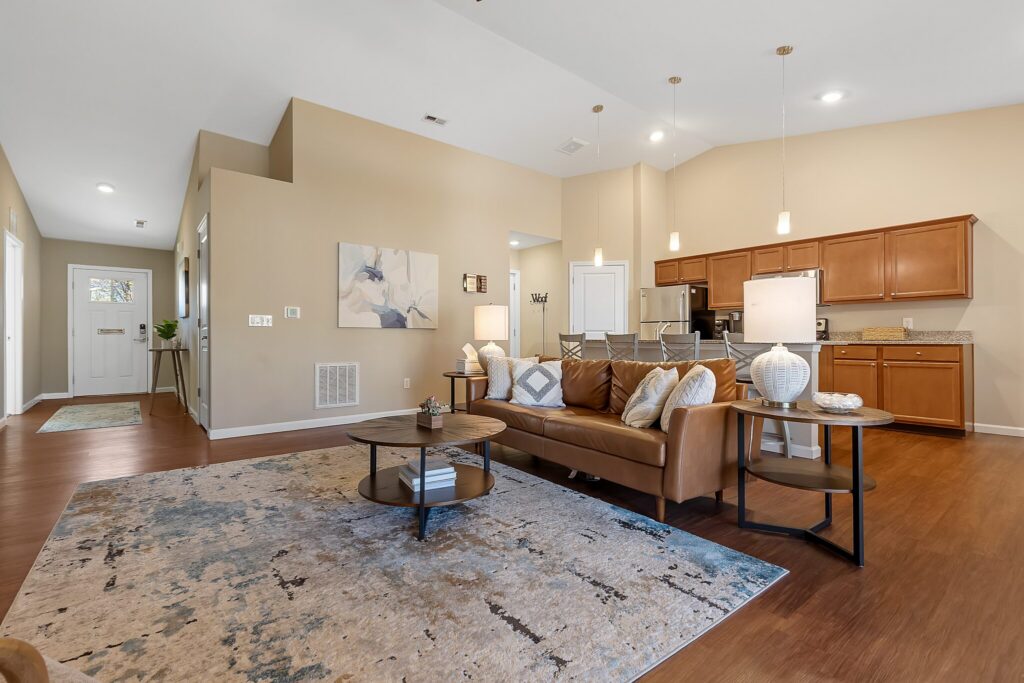 Open-concept living room with a brown leather sofa, round coffee table, area rug, and view of a kitchen with wooden cabinets and stainless steel appliances.