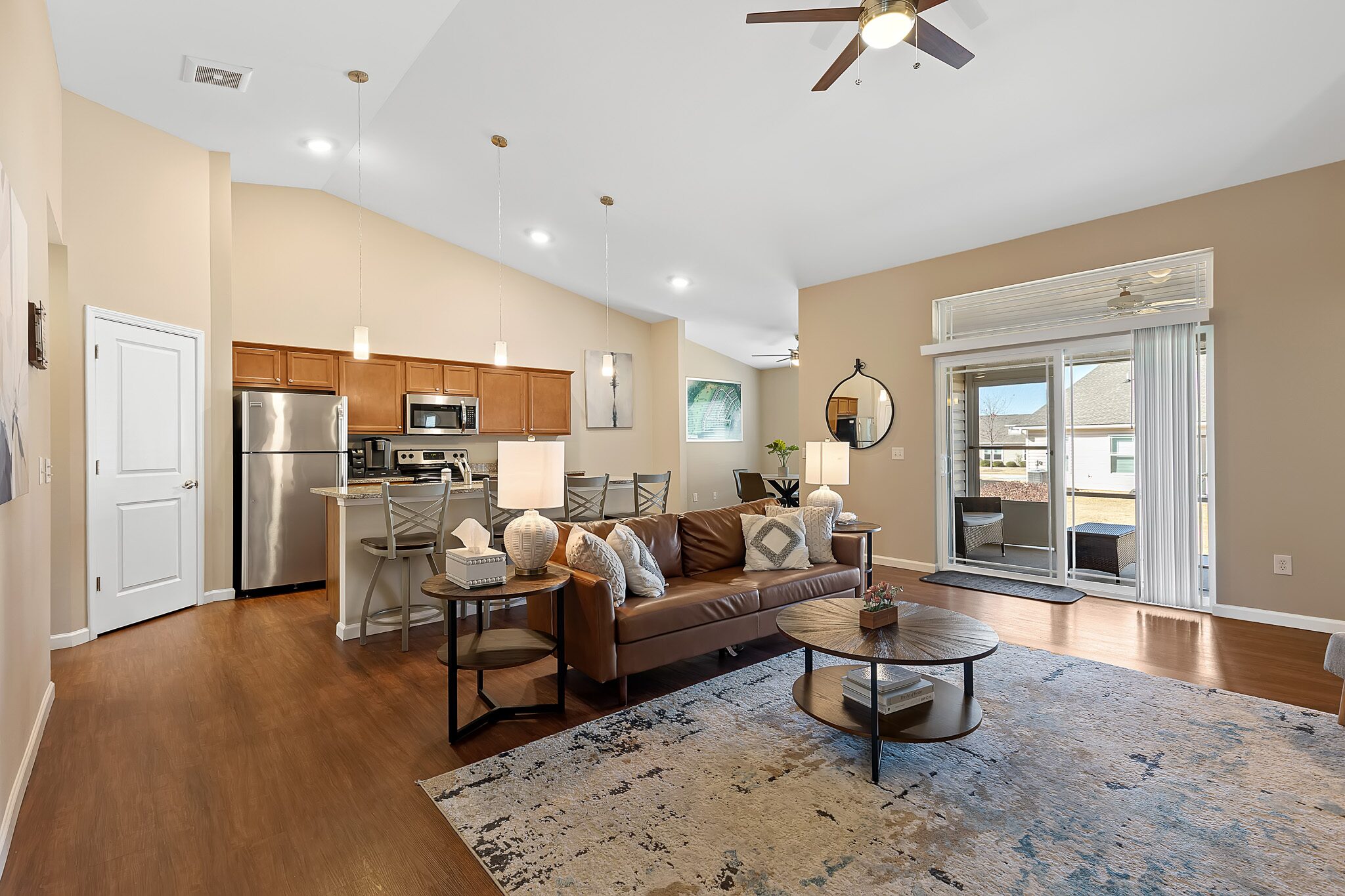 Open-concept living room and kitchen with brown cabinets, stainless steel appliances, leather sofa, round coffee table, and sliding glass doors leading to a patio.