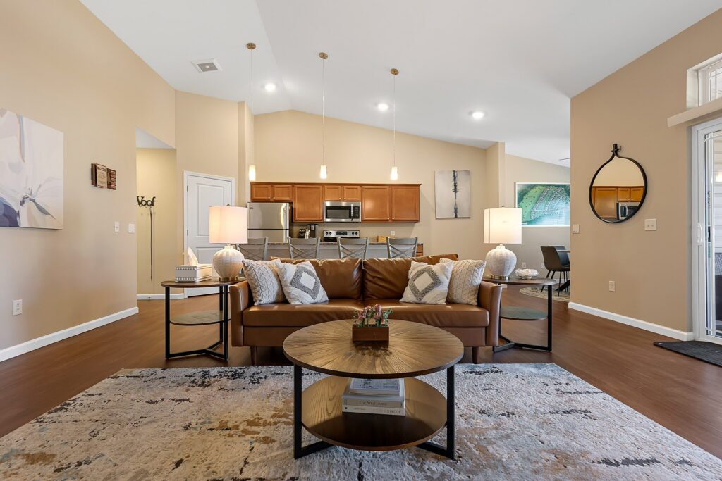 A modern living room with a brown leather sofa, round coffee table, area rug, and open view of a kitchen with wooden cabinets and stainless steel appliances.