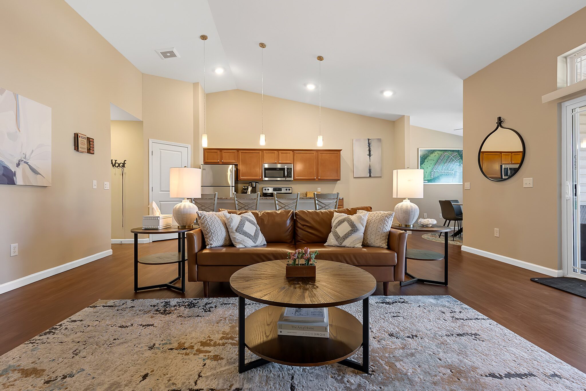 A modern living room with a brown leather sofa, round coffee table, area rug, and open view of a kitchen with wooden cabinets and stainless steel appliances.