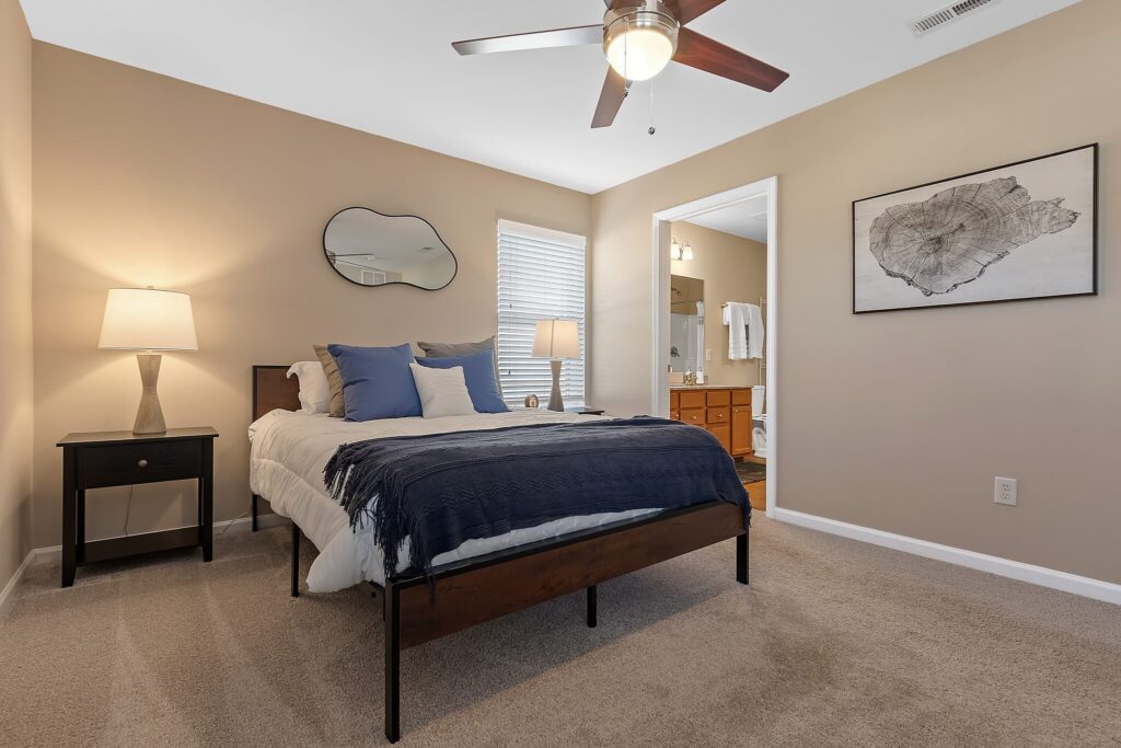 A neatly made bed with blue and white bedding, two bedside tables with lamps, a ceiling fan, beige walls, and an open doorway to a bathroom.