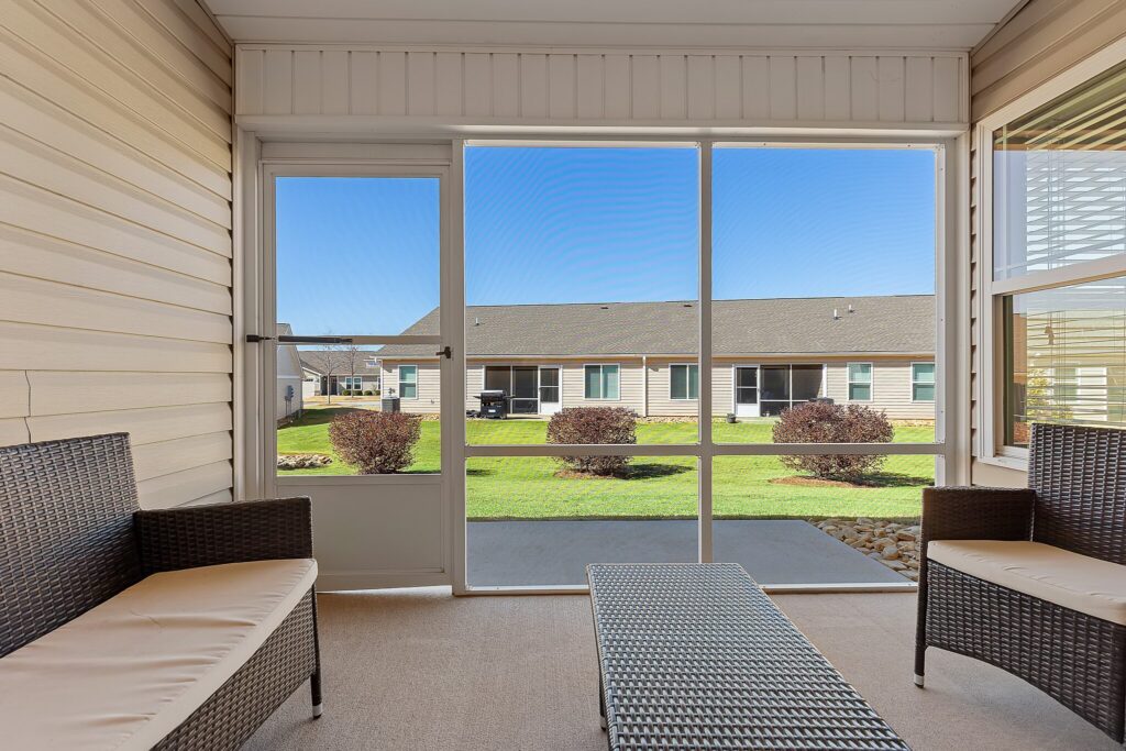 Screened-in porch with wicker furniture, overlooking a well-kept lawn and opposite building under a clear blue sky.