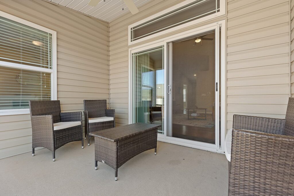 A covered patio with beige siding, two wicker armchairs, a wicker loveseat, a wicker coffee table, and a sliding glass door leading inside.