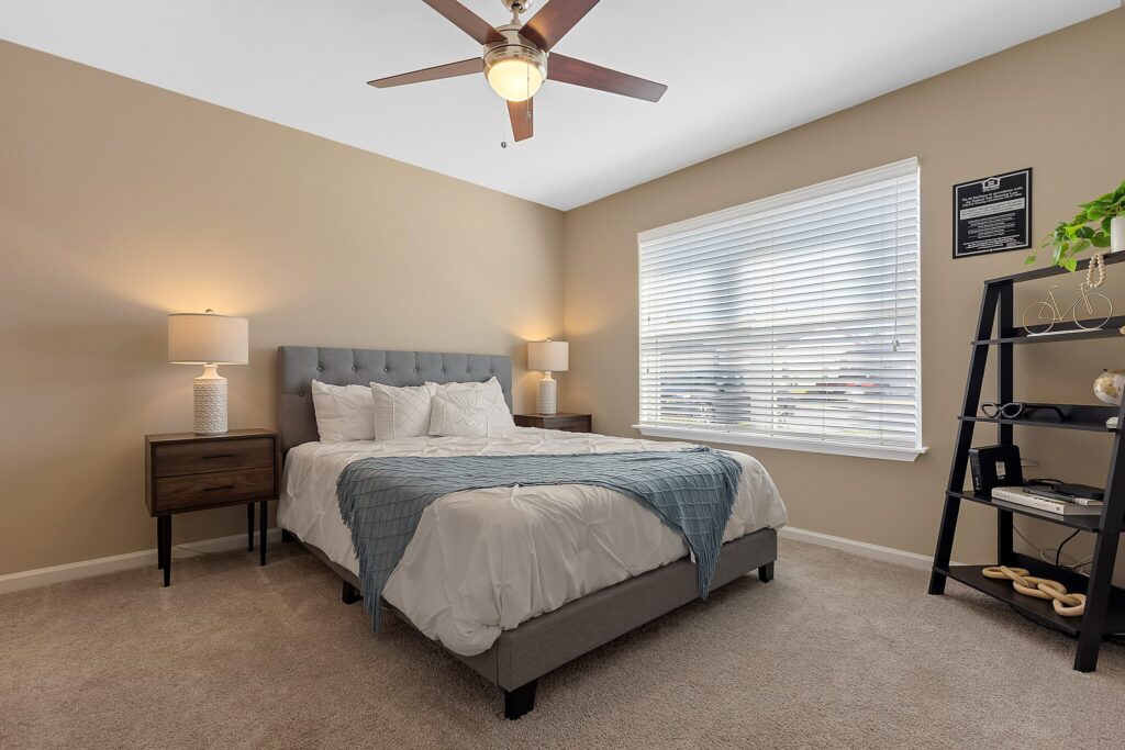 A modern bedroom with a gray upholstered bed, two bedside tables with lamps, a black shelf, beige walls, carpeted floor, and a ceiling fan. Natural light streams through a window.