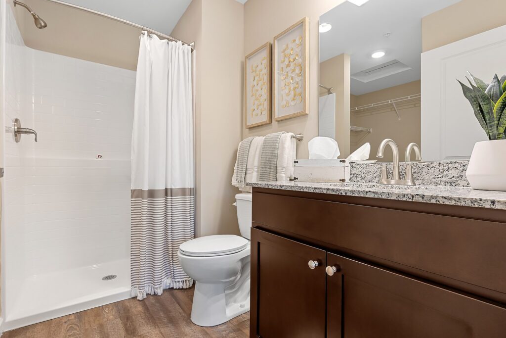 Modern bathroom with a shower, white curtain, toilet, dark wood vanity, granite countertop, towels, framed art, and a potted plant on the sink.
