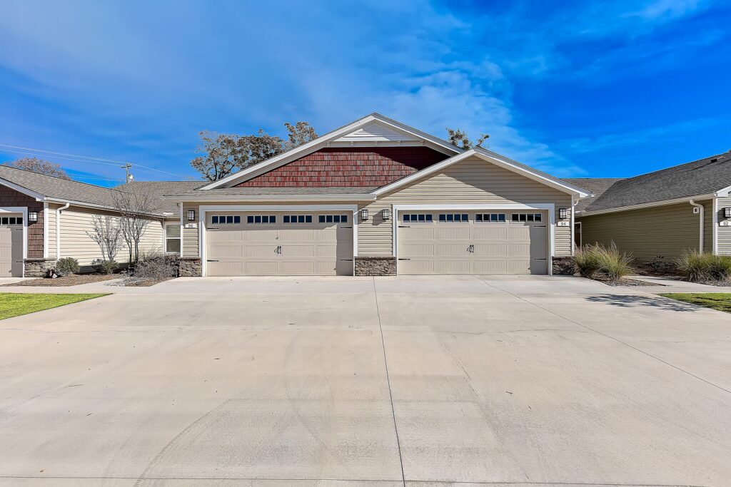 A duplex building with two double garage doors, beige siding, and a wide concrete driveway under a blue sky.