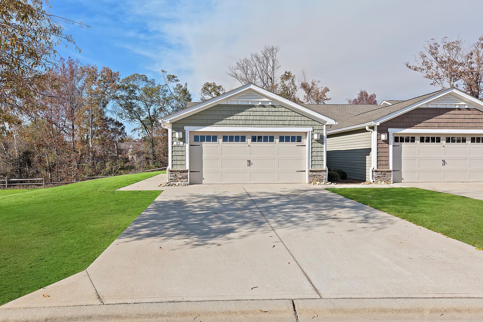 A duplex with two attached garages, each with light-colored doors and windows, sits on a neatly maintained lawn bordered by trees and a clear driveway.