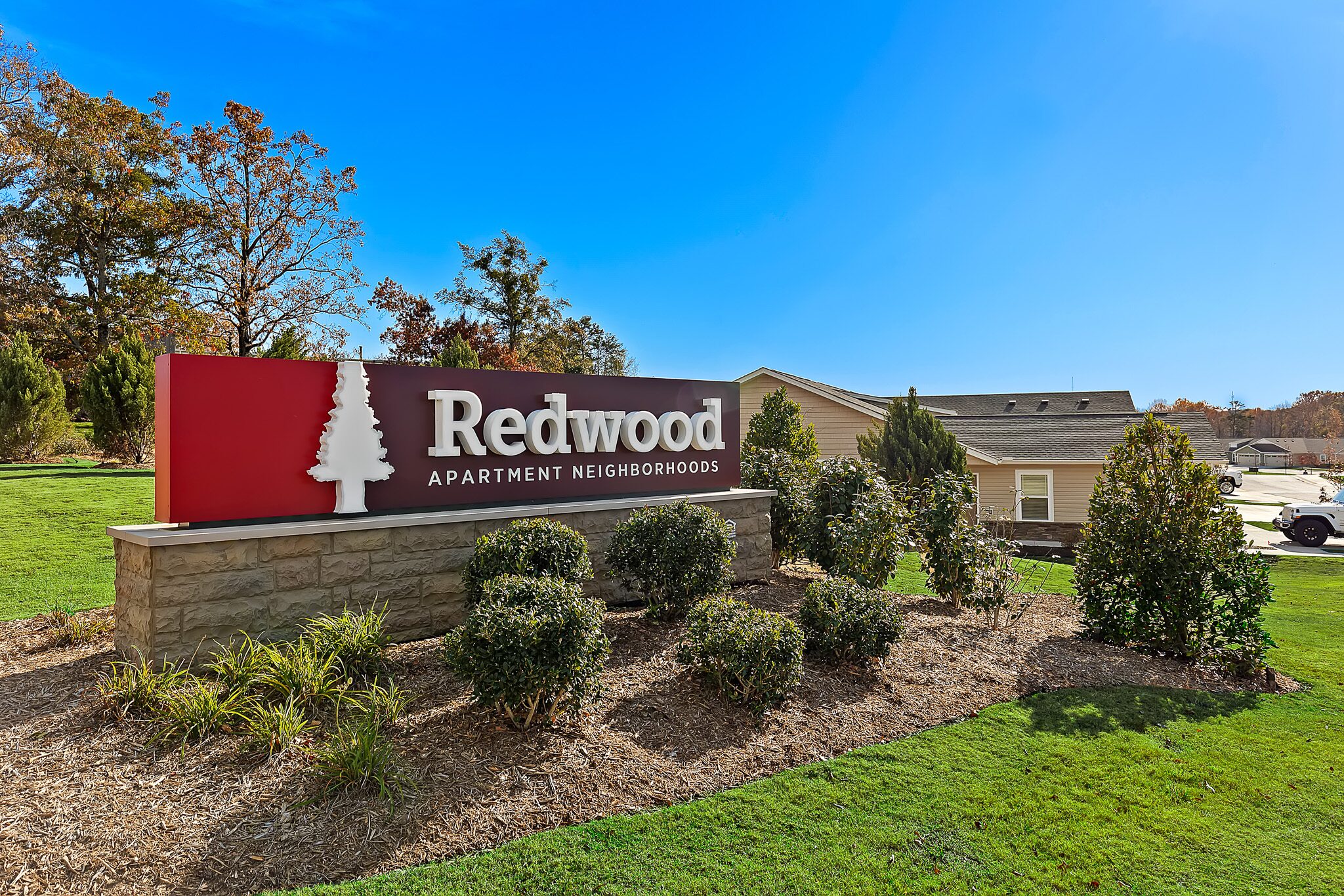 A sign for Redwood Apartment Neighborhoods stands on a landscaped lawn with bushes and a building in the background.
