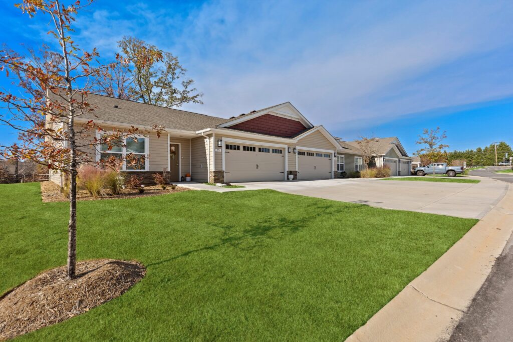 Single-story duplex homes with attached garages, a driveway, and neatly maintained lawns under a clear blue sky.