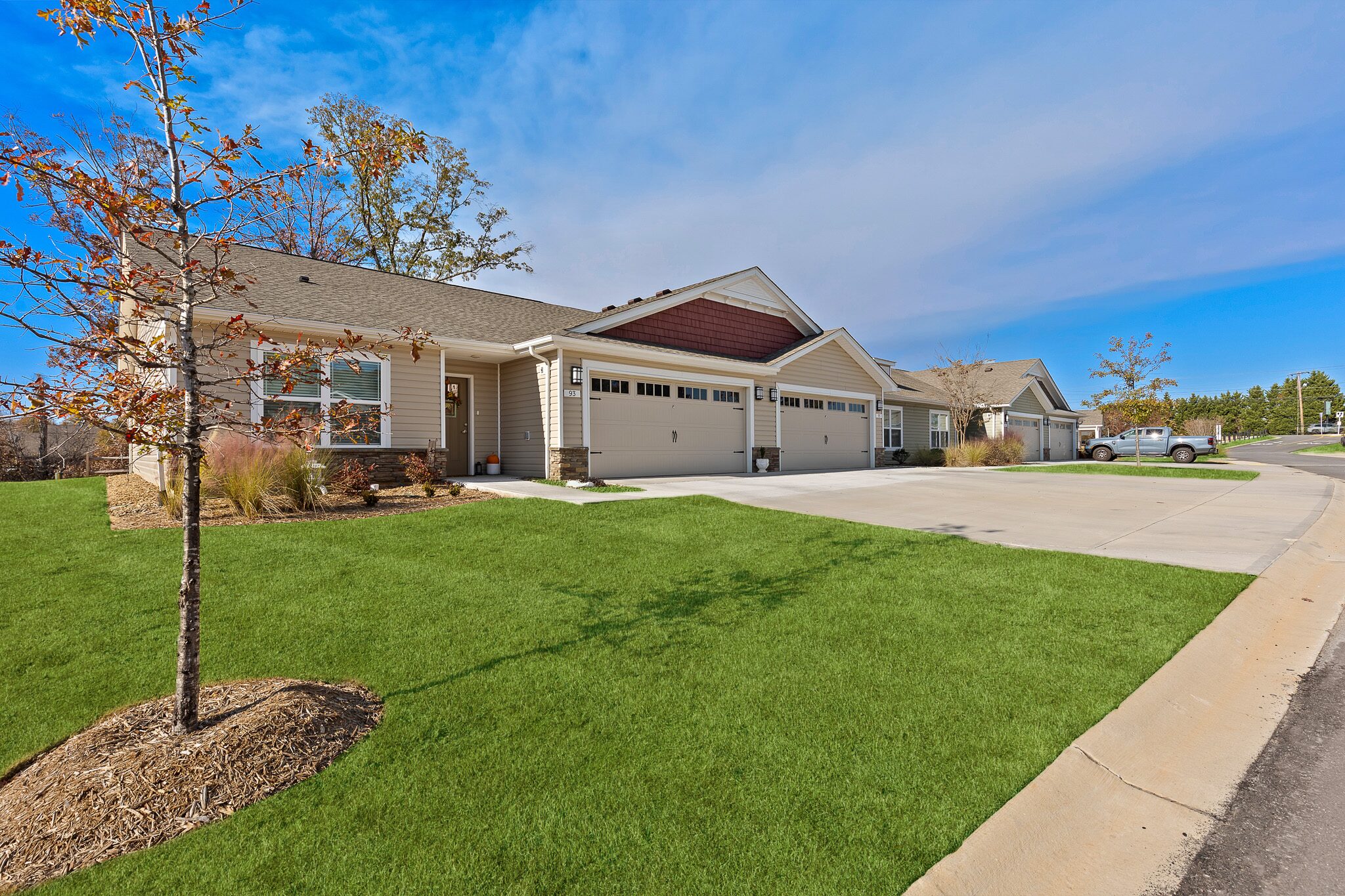 Single-story duplex homes with attached garages, a driveway, and neatly maintained lawns under a clear blue sky.