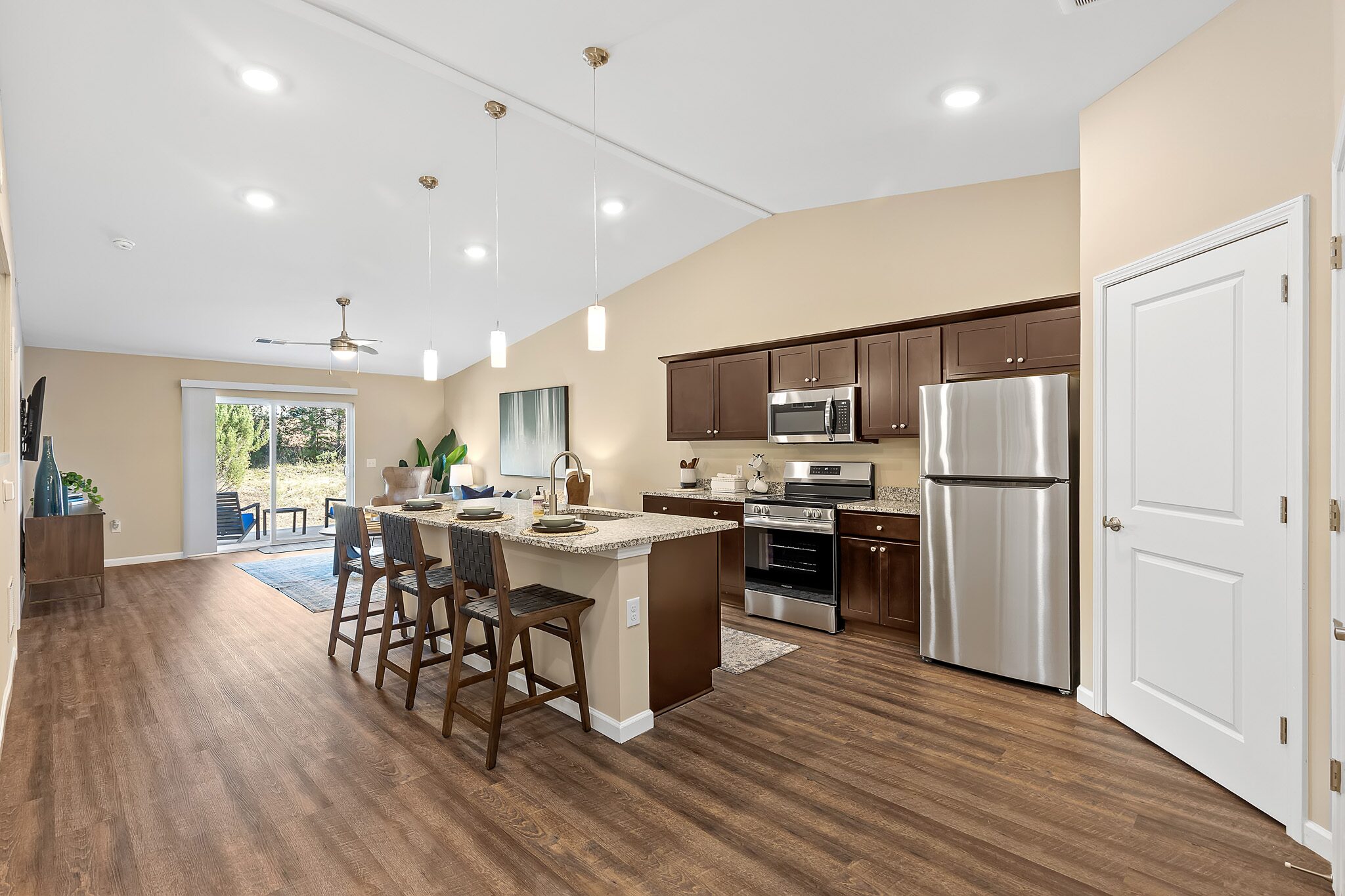 Modern kitchen with stainless steel appliances, dark wood cabinets, island with barstools, and open layout leading to living area with large windows.