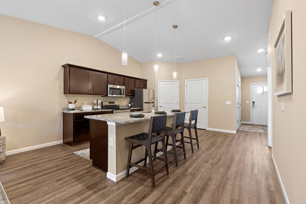 Modern kitchen with dark cabinets, stainless steel appliances, and a granite island with three chairs on wood flooring under pendant lighting.