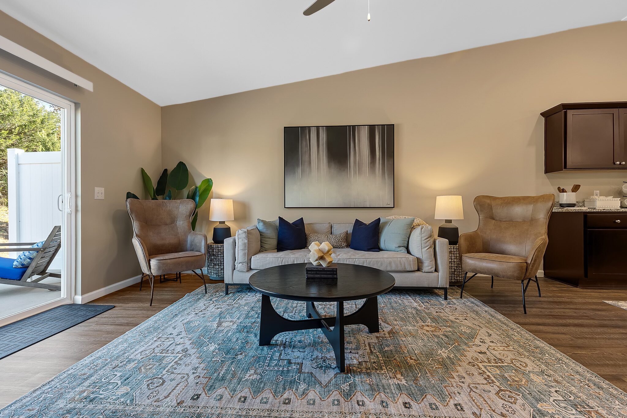 A modern living room with a beige sofa, two armchairs, a round black coffee table, a blue patterned rug, neutral walls, and sliding glass doors to an outdoor area.
