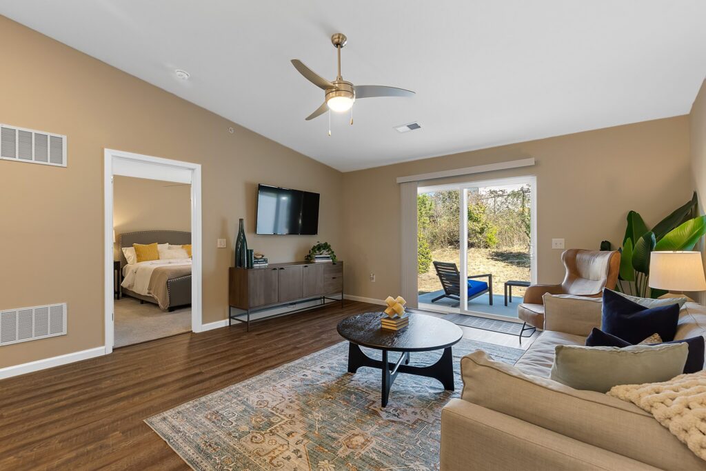 Spacious living room with a sofa, coffee table, TV, and large glass doors opening to a patio; bedroom visible through an open doorway.
