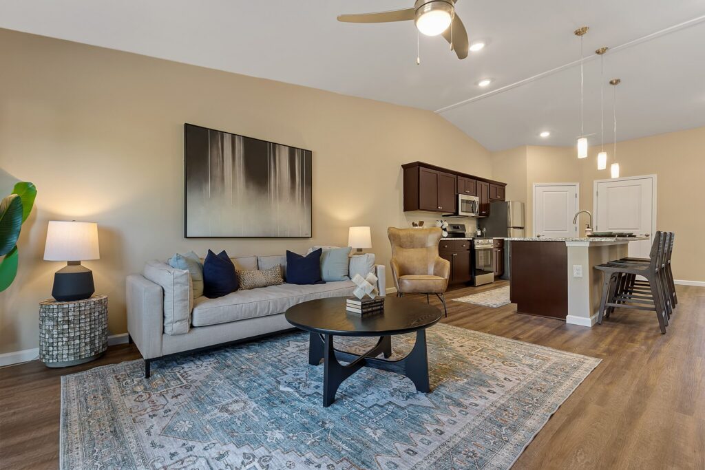 Modern open-concept living room and kitchen with beige walls, neutral furniture, a large rug, pendant lights, and stainless steel appliances.