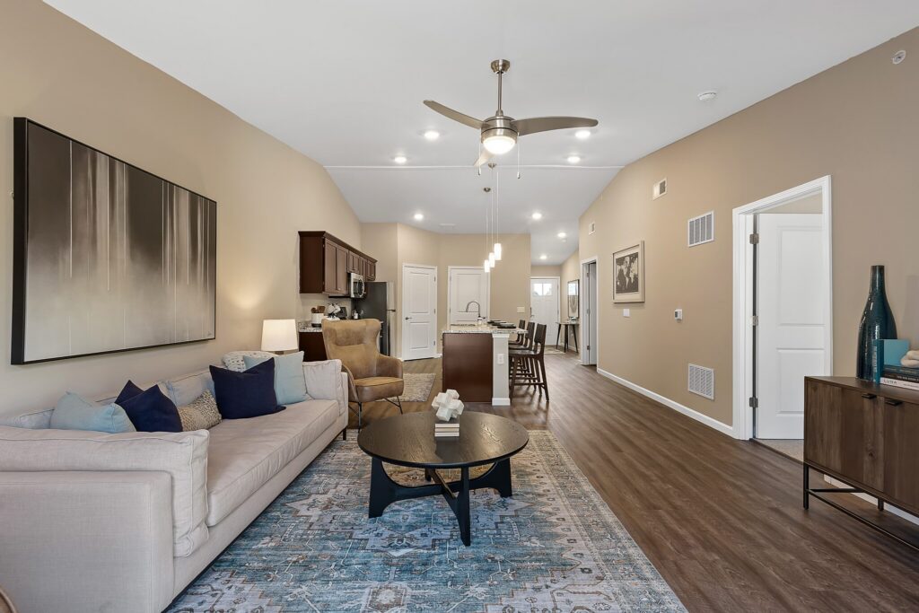 Modern open-concept living room and kitchen with neutral walls, wooden flooring, beige sofa, blue accents, round coffee table, and ceiling fan.