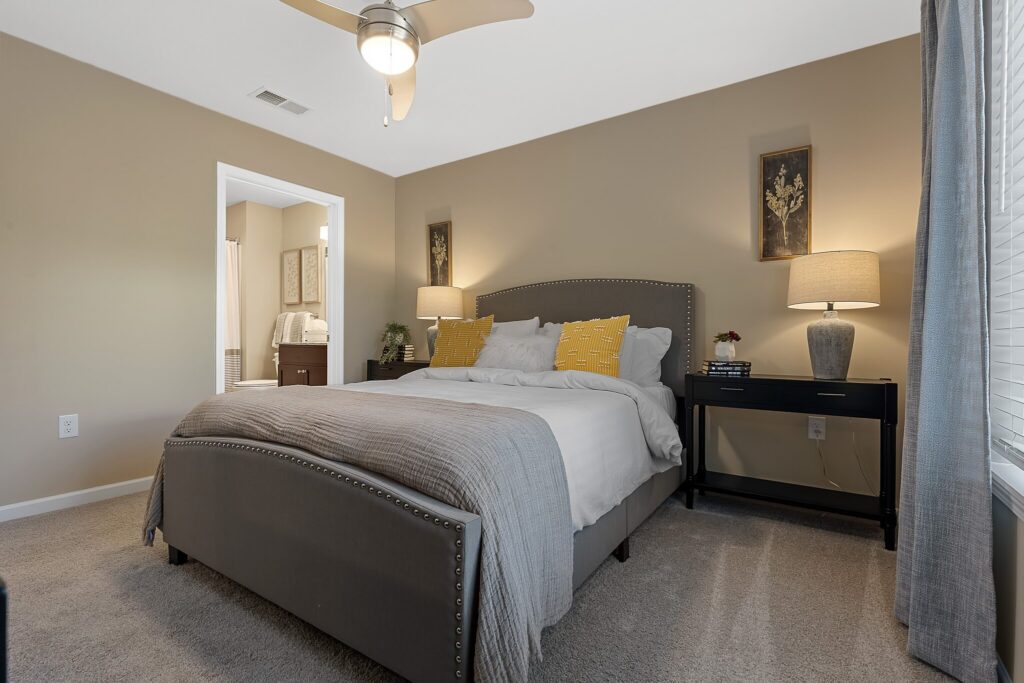 A neatly made bed with gray upholstery and yellow pillows sits between two black nightstands with lamps in a beige-walled bedroom. An open door leads to a bathroom.