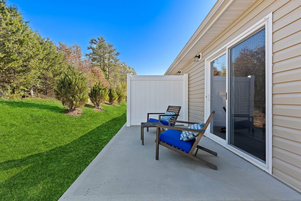 A concrete patio with two chairs and a small table, backed by a privacy fence and sliding glass doors, overlooks a grassy yard with trees.