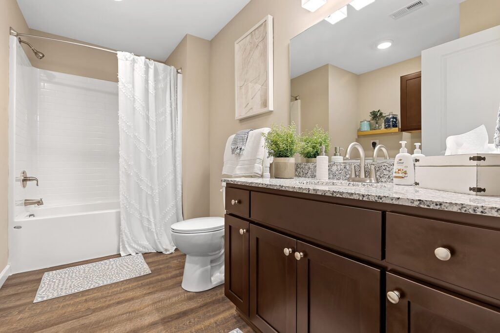 Modern bathroom with dark wood cabinets, granite countertop, white fixtures, a bathtub with a patterned shower curtain, and decorative plants on the vanity.