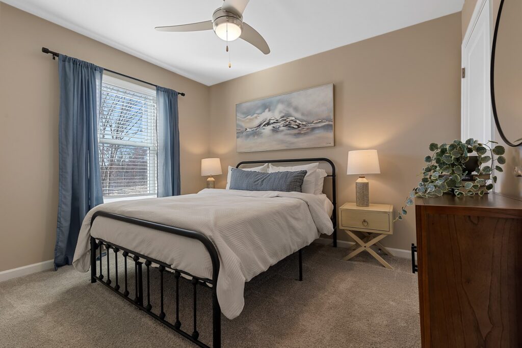 A neatly made bed with white bedding in a beige-walled bedroom, featuring two bedside tables with lamps, a dresser, and a window with blue curtains.
