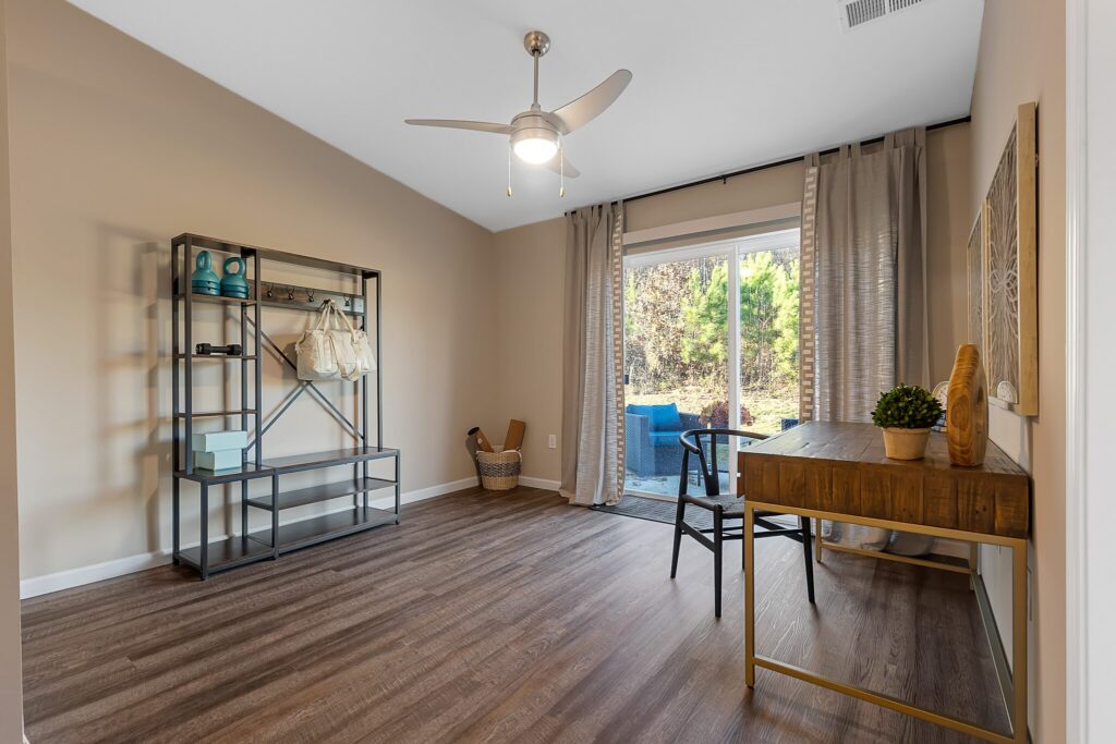 A home office with wood flooring, a metal shelving unit, a wooden desk with a black chair, floor-length curtains, and a glass door leading to an outdoor patio area.