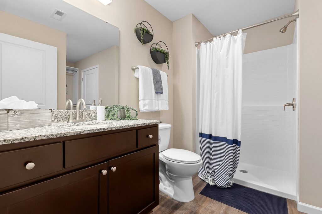 Modern bathroom with a granite countertop, dark wood vanity, white toilet, shower with a striped curtain, wall shelves with plants, and blue-striped floor mat.
