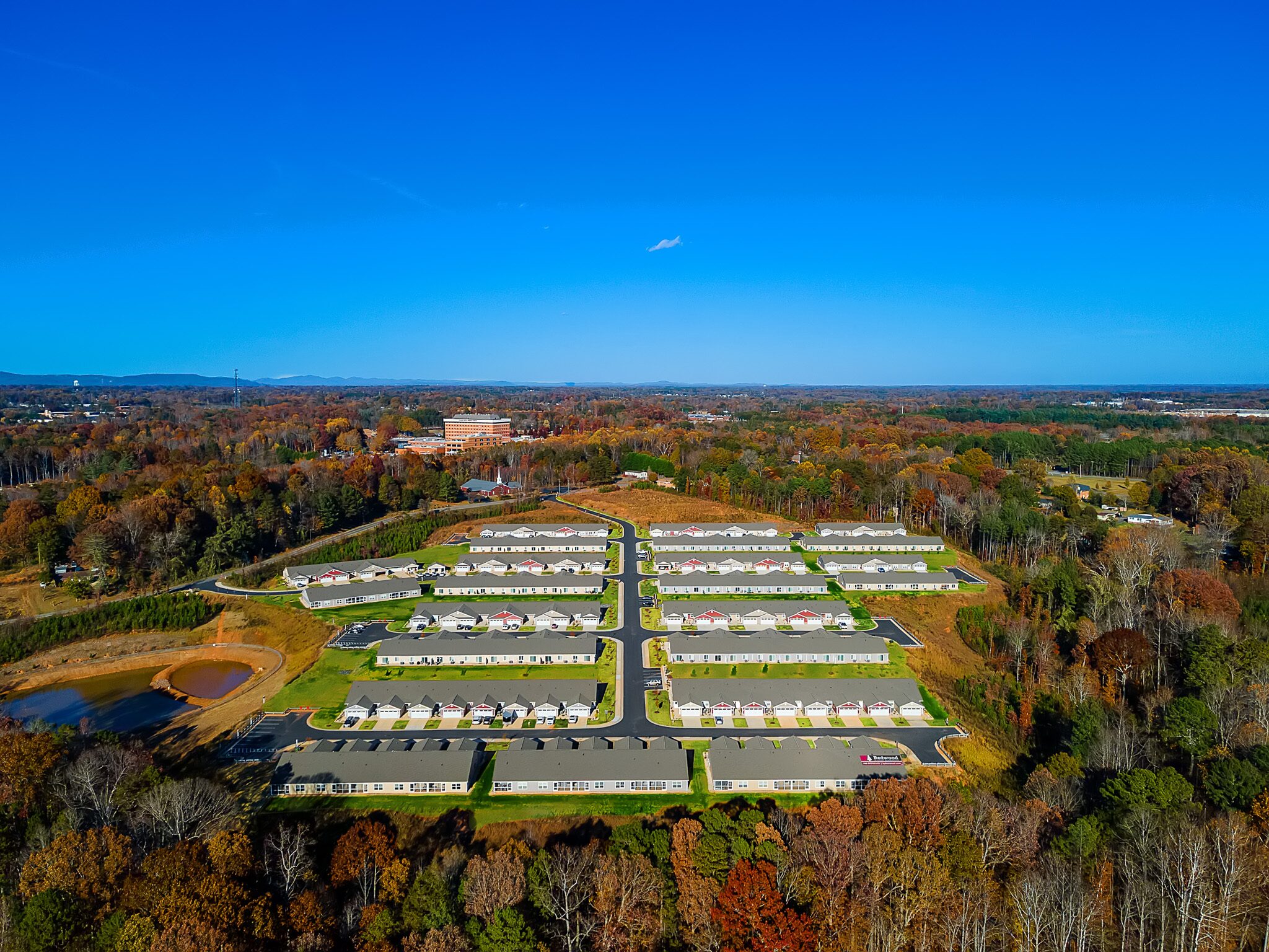 Aerial view of a residential neighborhood with rows of single-story houses, surrounded by trees and roads, under a clear blue sky.