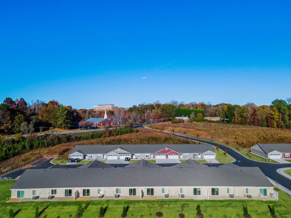 Aerial view of single-story residential buildings with lawns in front, a winding road, and a wooded hillside with autumn foliage in the background under a clear blue sky.