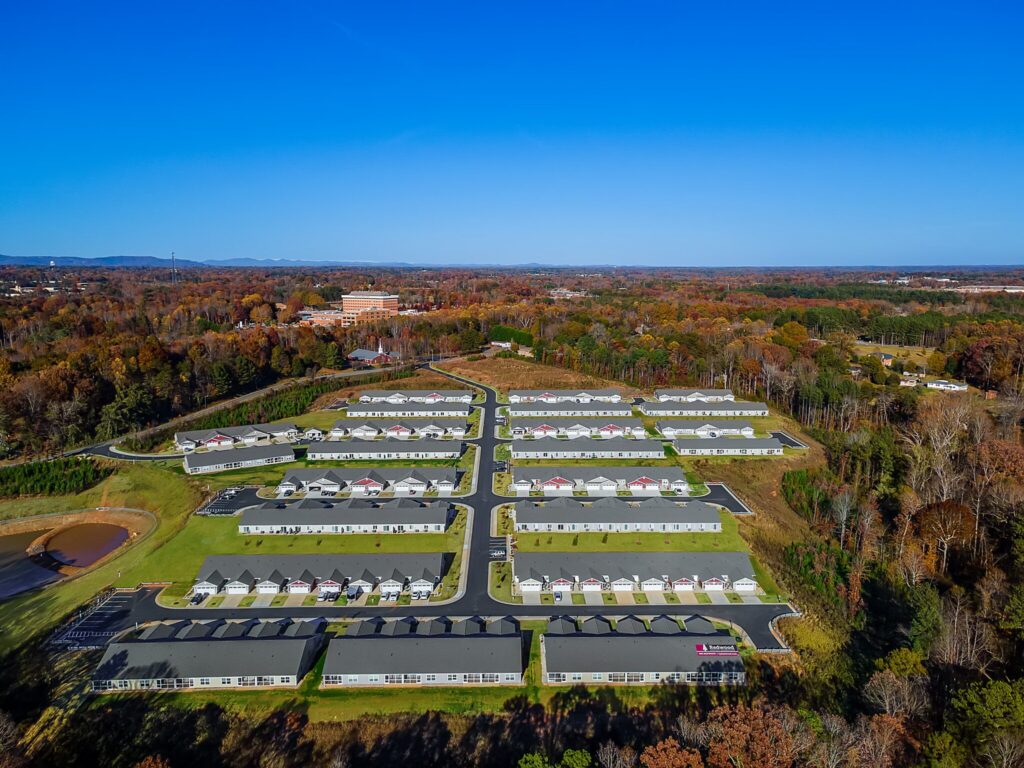 Aerial view of a suburban residential neighborhood with rows of houses, green lawns, and surrounding trees under a clear blue sky.